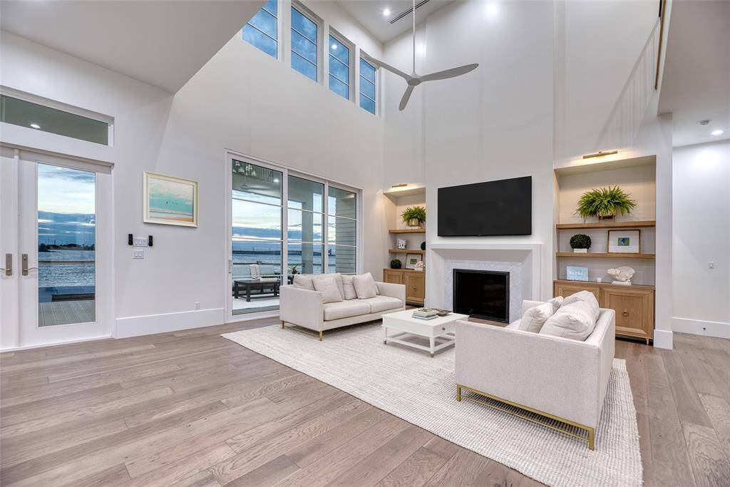 This spacious living room features high ceilings, a modern fireplace, and large windows offering stunning waterfront views. It includes elegant built-in shelves, a sleek ceiling fan, and opens to a patio, blending indoor and outdoor living beautifully.