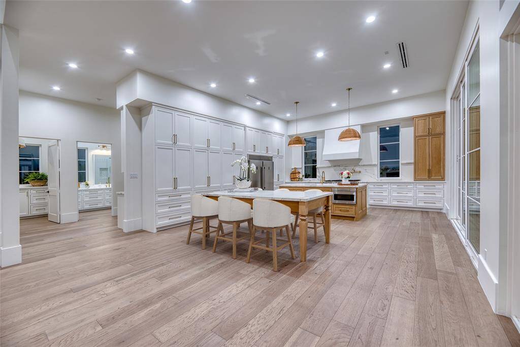 This spacious, modern kitchen features sleek white cabinetry, a large island with seating, and warm wood accents. It boasts ample natural light from large windows and elegant pendant lighting, perfect for cooking and entertaining.