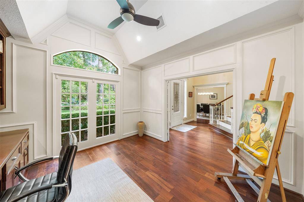 This bright home office features high ceilings, elegant wood flooring, and ample natural light from French doors. It includes built-in shelves, a ceiling fan, and has an open view into the dining area, creating a welcoming and functional workspace.