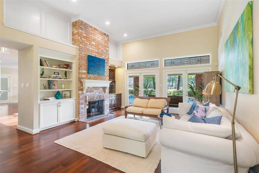 Bright and spacious living room with high ceilings and large windows. Features a cozy brick fireplace, built-in shelving, and hardwood floors. Ample natural light and a view of the golf course create a welcoming atmosphere.