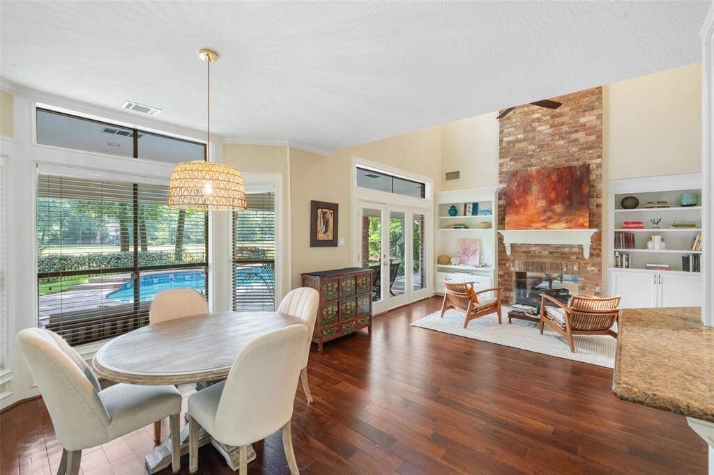 This bright, open-concept living area features hardwood floors, a cozy fireplace with built-in shelves, and large windows offering views of the golf course and pool. The space includes a breakfast area with modern lighting and an inviting atmosphere.