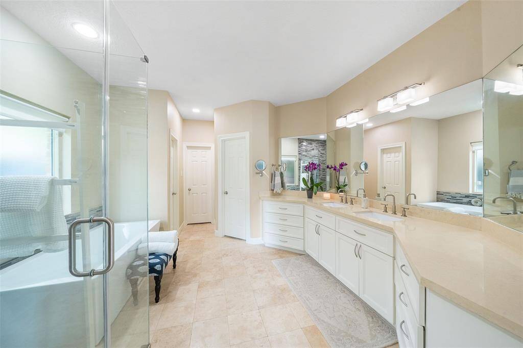 This spacious bathroom features a modern design with dual sinks, ample countertop space, and a large mirror. It includes a glass-enclosed shower, a separate bathtub, and neutral tiling. Natural light brightens the room, enhancing its airy feel.