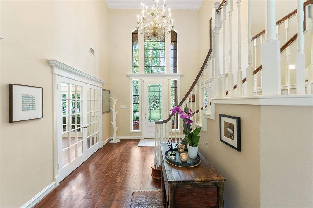This elegant foyer features high ceilings, a grand staircase, and rich hardwood floors. A large window overlooking the lush front yard allowing natural light to flood the space, enhanced by a stunning chandelier. French doors lead to a study with built-in bookshelves, adding a touch of sophistication.
