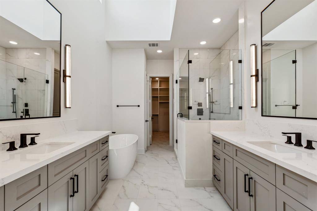 Indulge in a spa-like experience in the primary bath, featuring soaring ceilings, ample natural light from a large window, and double custom-built vanities with Kohler undermount sinks. Visual Comfort sconces frame the mirrors, and sleek porcelain tile floors create a chic, modern look. Straight ahead the door opens to a huge walk-in closet with his and hers dressers and hidden storage that completes the ultimate retreat.