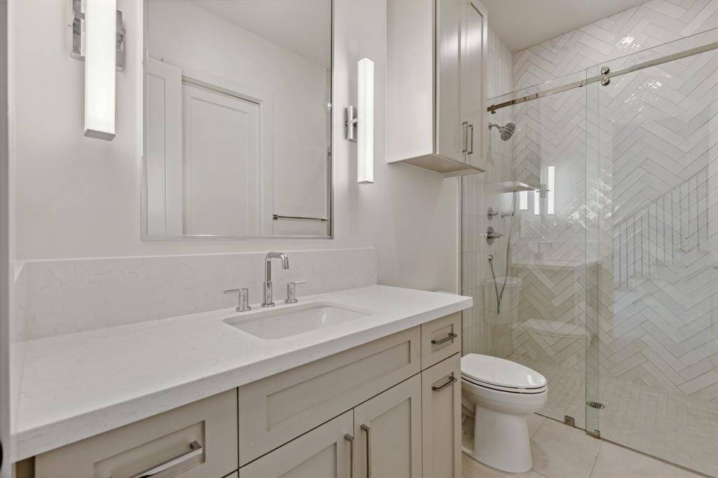 This sleek en-suite bathroom for secondary bedroom #2 features a custom-built vanity with ample storage, Top Knob hardware, and a quartz countertop with an 8-inch backsplash. A framed metal mirror adds a modern touch, while the walk-in shower with herringbone tile, soap niche, and water-repellent sliding glass door elevates the space. Kohler toilet and 24x48 tile flooring complete the clean, contemporary design.