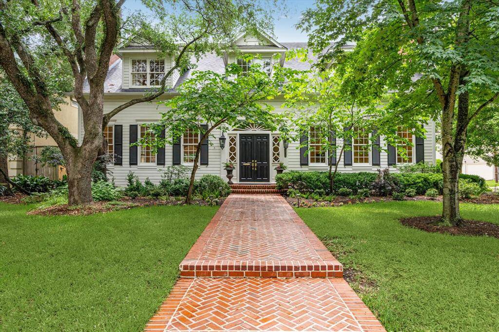 A stately brick walkway leads to this classic Colonial facade, framed by lush landscaping and mature trees. The symmetrical design, black shutters, and grand double doors evoke timeless elegance.