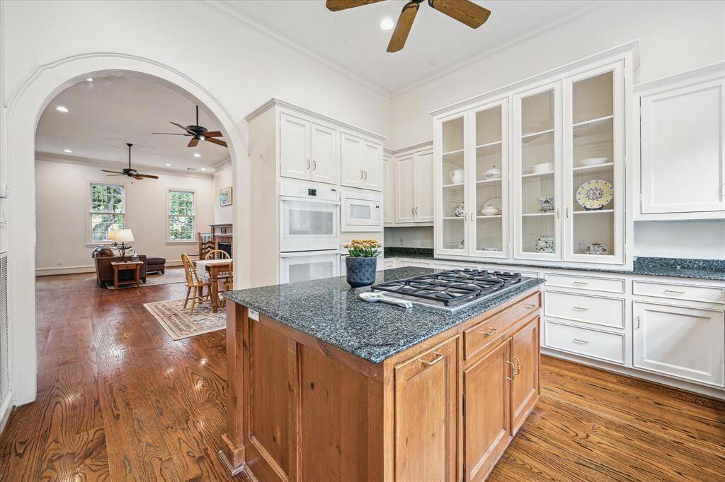 This expanded view showcases the kitchen’s harmonious blend of style and function, featuring a spacious island with gas cooktop, double ovens, abundant cabinetry with glass display fronts, and a graceful archway that connects seamlessly to the adjoining living space.