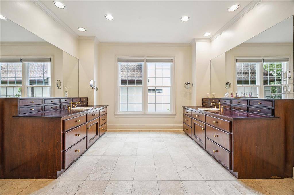 The primary bathroom features dual vanities with rich wood cabinetry, elegant brass fixtures, and full-wall mirrors, all bathed in natural light from oversized windows.