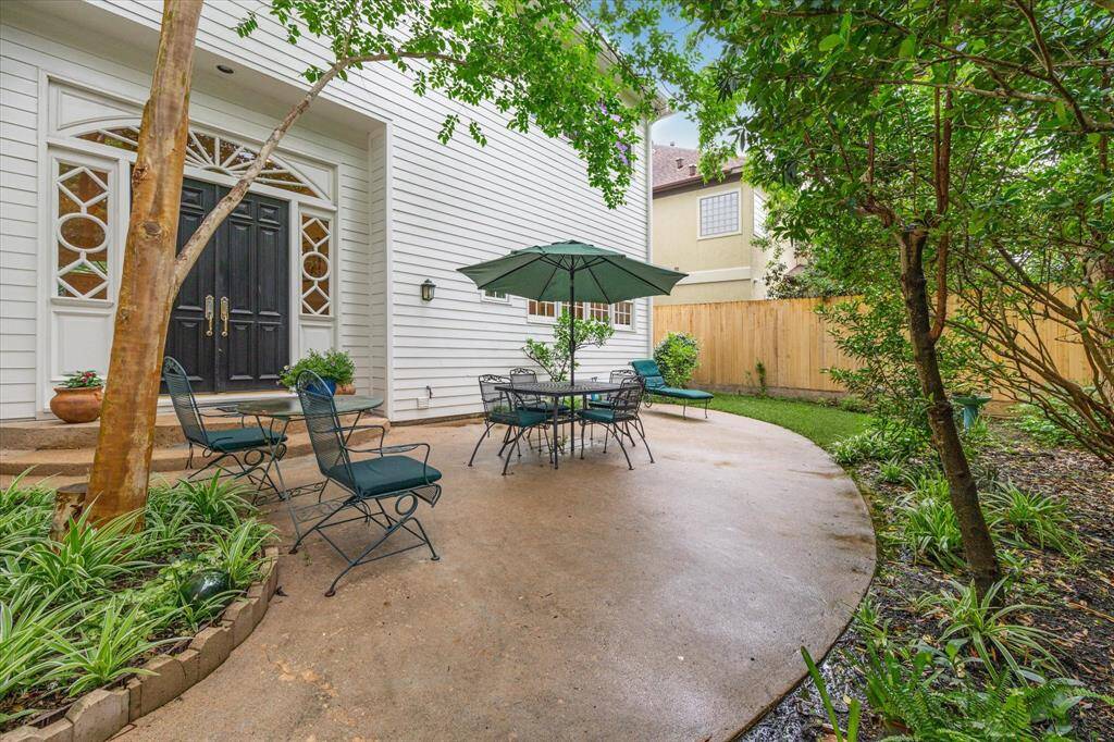 This additional patio view highlights the inviting double-door entry, lush surrounding greenery, and ample space for outdoor dining and lounging beneath the shade