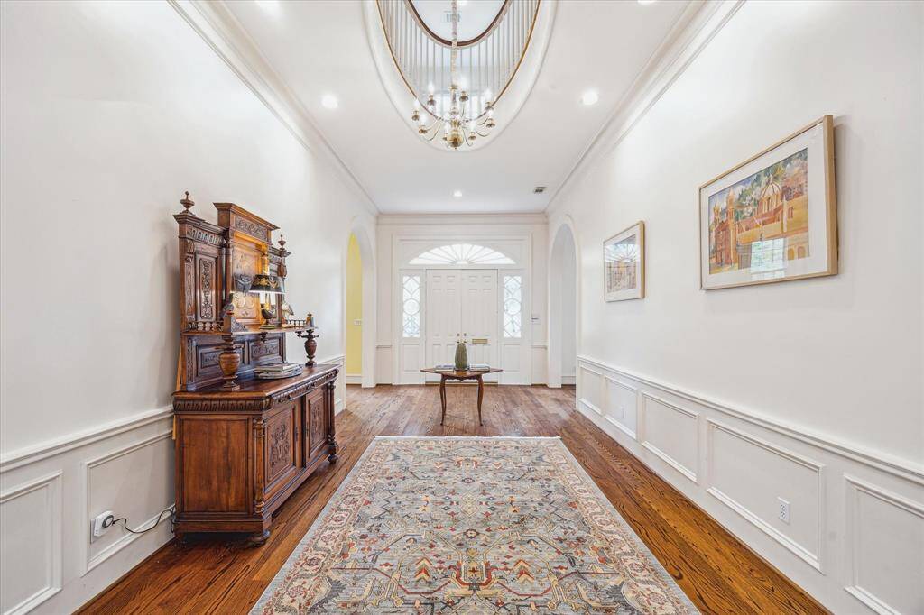 The grand entryway impresses with rich hardwood floors, elegant wainscoting, and a soaring oval staircase opening to a chandelier-lit ceiling above.