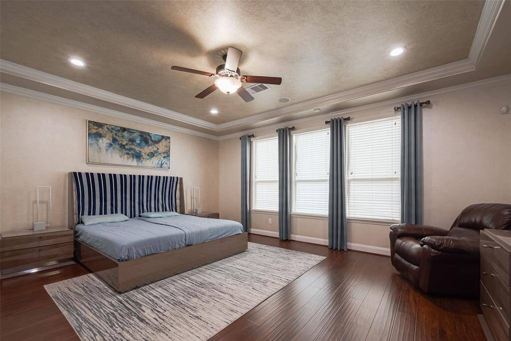 Spacious primary bedroom with elegant tray ceiling and recessed lighting. Features include a modern ceiling fan, large windows with blue curtains, wood flooring, and a cozy seating area. The room is tastefully decorated with a contemporary bed and artwork, creating a welcoming and serene atmosphere.