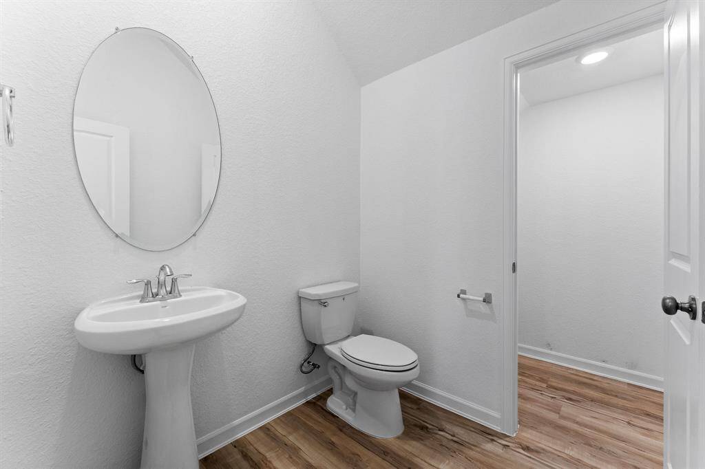 Half bath for guests features a pedestal sink,  wood-look laminate flooring, toilet, and an oval mirror.There is also a large room off this area for storage.