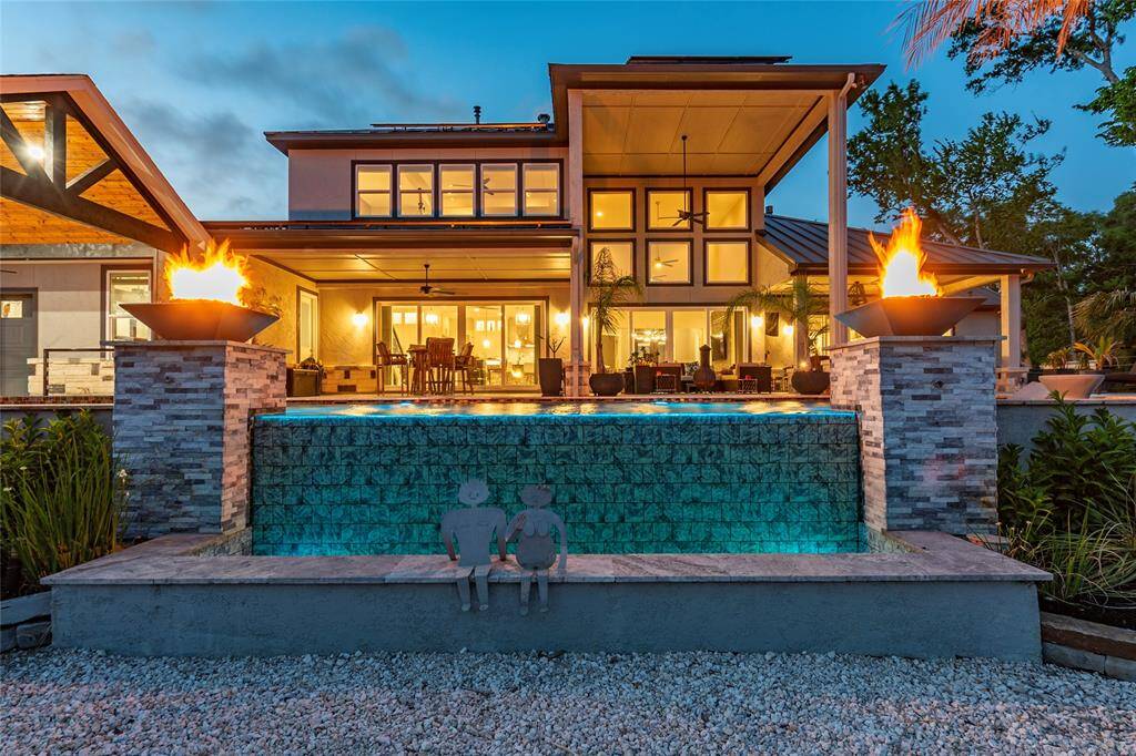 A glowing infinity-edge water wall cascades gracefully into a tiled catch basin, creating a mesmerizing focal point accented by dramatic fire-water bowls. Framed by dual 18" x 30" tile columns topped with elegant travertine caps, this striking feature brings resort-style ambiance and architectural sophistication to the outdoor oasis.