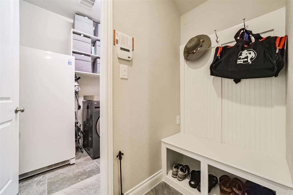 his photo showcases a tidy mudroom and laundry area. It features a bench with storage cubbies underneath and hooks above for hanging items. The space includes a washing machine, shelving for organization, and a large freezer. The neutral color palette creates a clean and functional look.