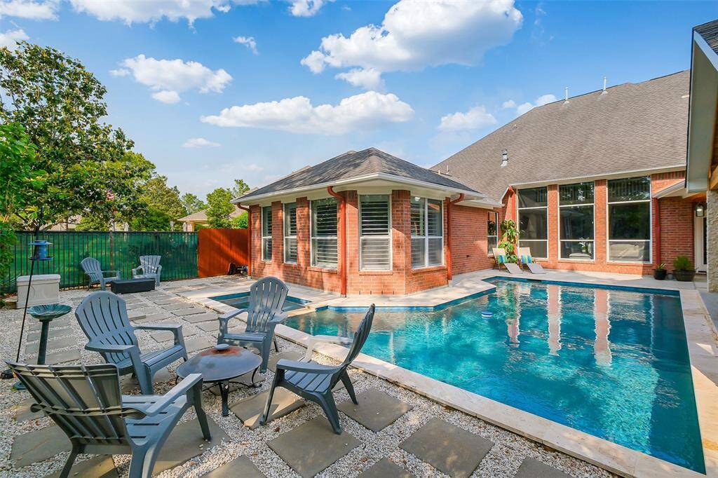 A sleek, in-ground pool with clean geometric lines, surrounded by travertine stone decking. The water shimmers under ambient LED lighting, reflecting the silhouettes of swaying trees and the soft glow from the nearby fire pit.