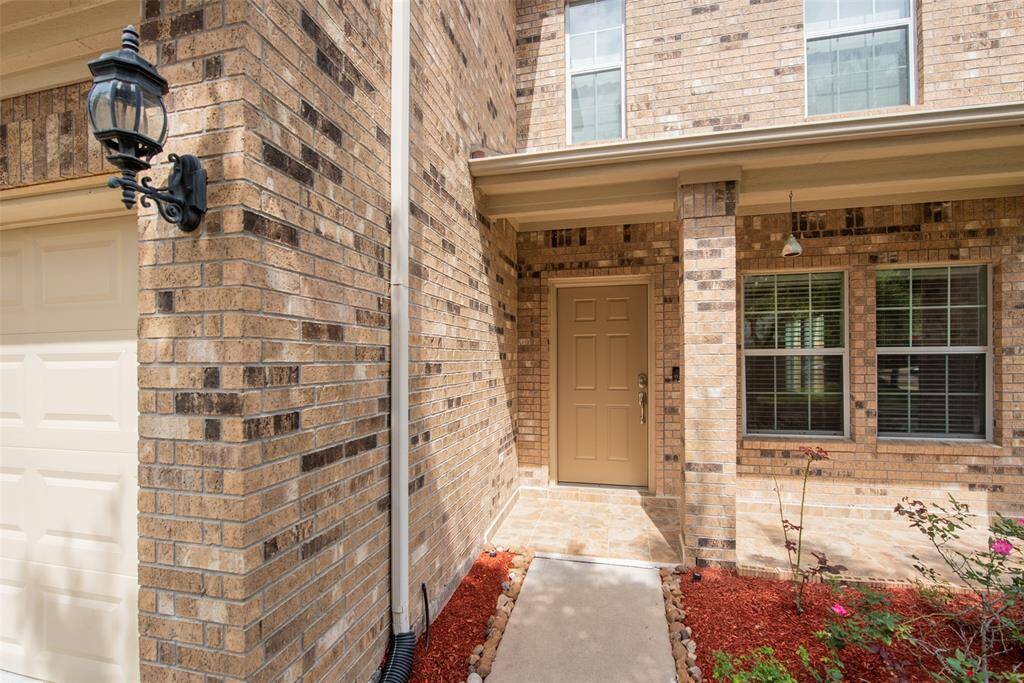 This photo showcases a welcoming front entrance of a brick home with a neutral-toned door, elegant porch lighting, and a well-maintained garden area with mulch and plants.