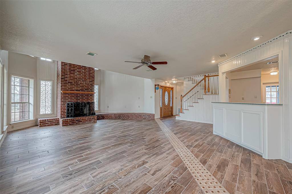 A dramatic floor-to-ceiling brick accent wall commands attention, housing a striking fireplace that serves as the room’s focal point. The soaring ceiling adds grandeur, while large windows flood the space with daylight.