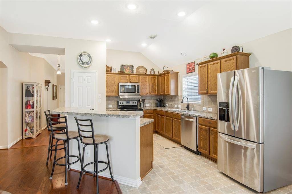 This open kitchen features warm wooden cabinets, sleek stainless steel appliances, and a convenient breakfast bar with seating. Tiled flooring and ample natural light enhance the bright, welcoming atmosphere.