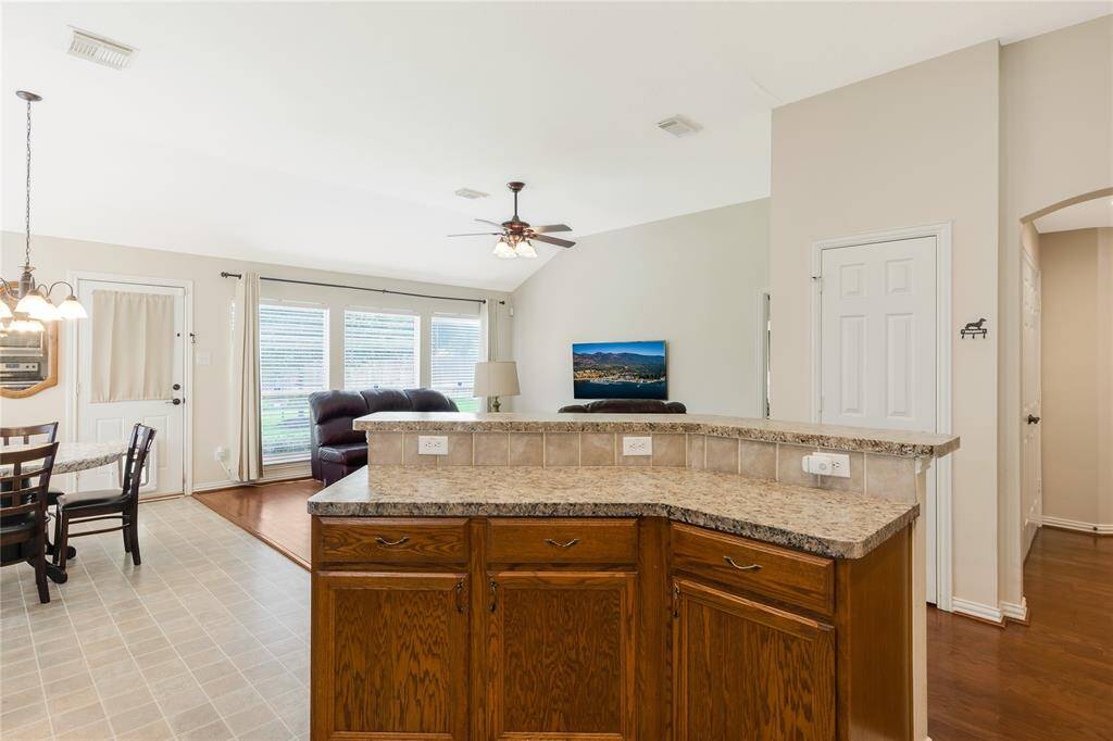Spacious kitchen with a large island with seating, overlooks a cozy living room with abundant natural light from the multiple windows. Adjacent to the space is a charming dining area with a chandelier, all tied together by neutral walls and warm wood flooring for a cohesive, inviting feel.