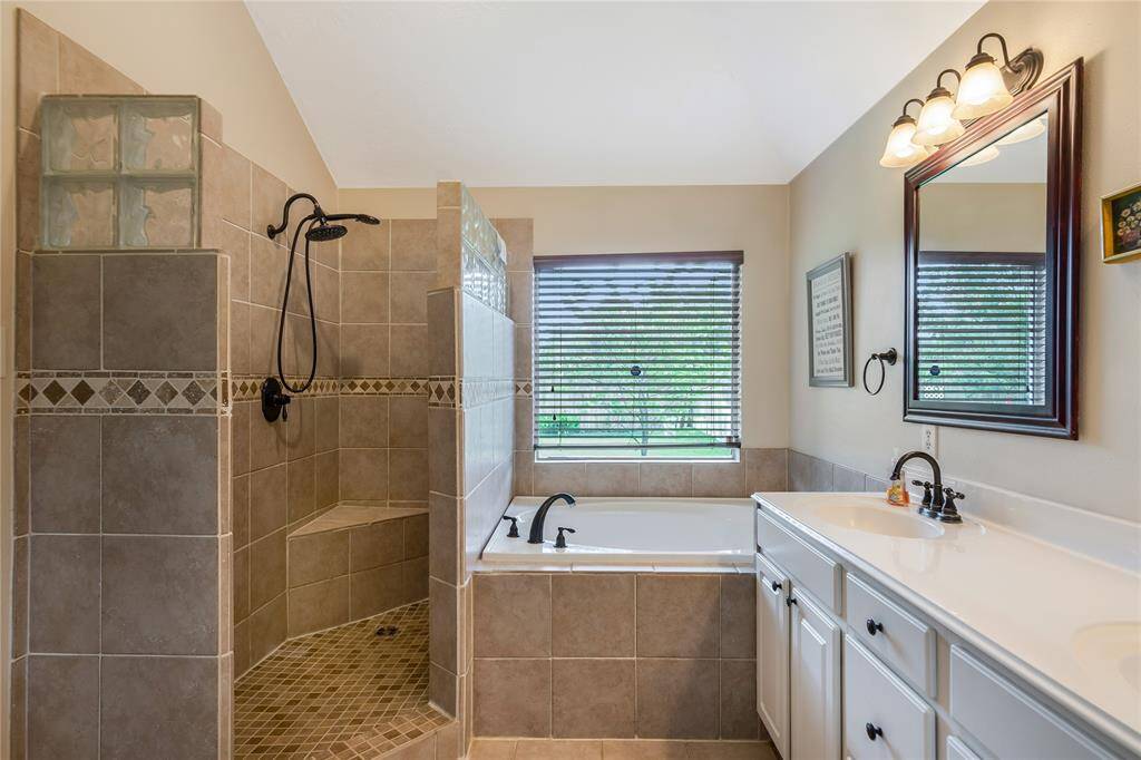 This bathroom features a spacious walk-in shower with decorative tiles, a separate bathtub under a window with blinds, and a double-sink vanity with a large mirror and elegant lighting, creating a bright and inviting space.