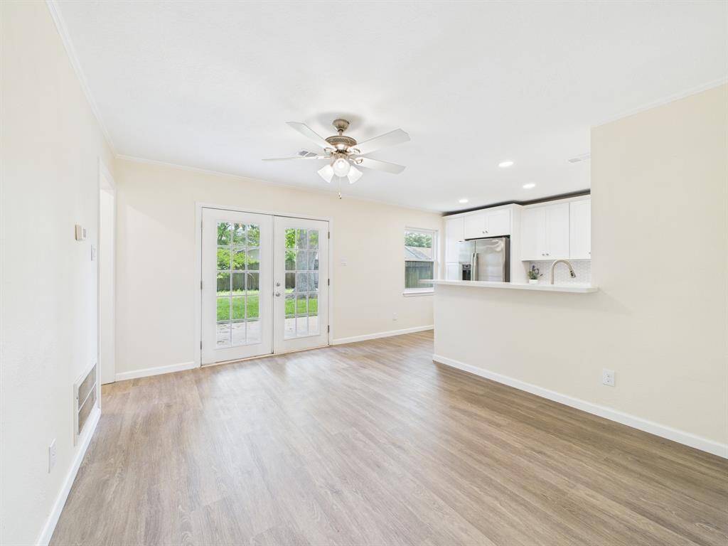 Bright and airy open-plan living area with wood flooring, a ceiling fan, and French doors leading to a backyard. The adjoining kitchen features modern white cabinetry, stainless steel appliances, and a breakfast bar.