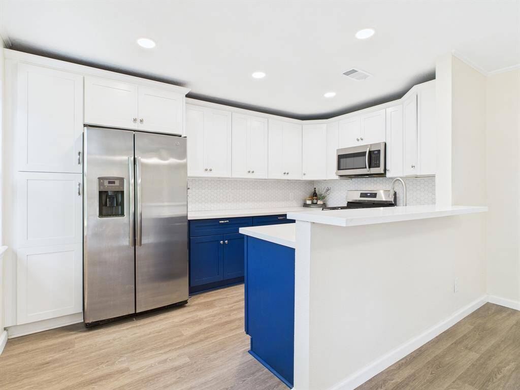 This modern kitchen features white cabinets with a striking blue accent, stainless steel appliances, and a sleek countertop. The open layout and wood-like flooring provide a spacious and inviting feel.