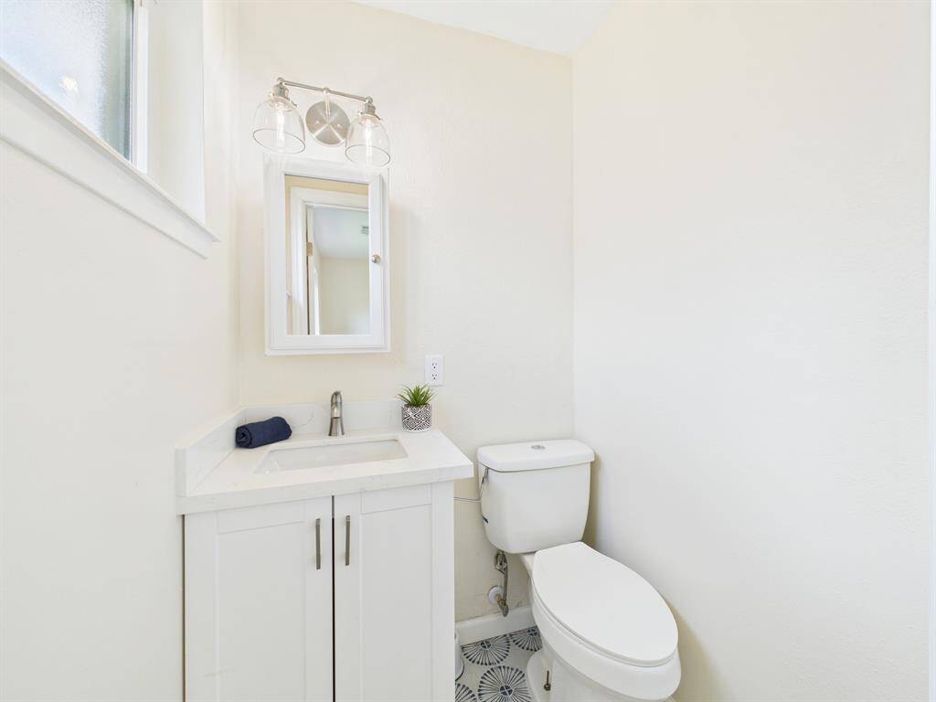 This primary 1/2 bathroom is a bright, compact bathroom featuring a white vanity with a mirror, a modern light fixture, and a toilet. The space is accented by light walls and a small window, creating an airy feel.