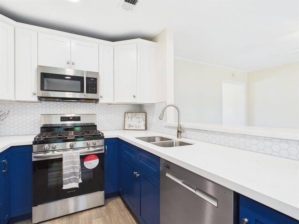 Modern kitchen with a sleek design featuring white upper cabinets and blue lower cabinets. It has stainless steel appliances, including a gas stove, microwave, and dishwasher. The countertops are white with a hexagonal tile backsplash, and there's a double sink with a contemporary faucet. The space is bright and inviting.