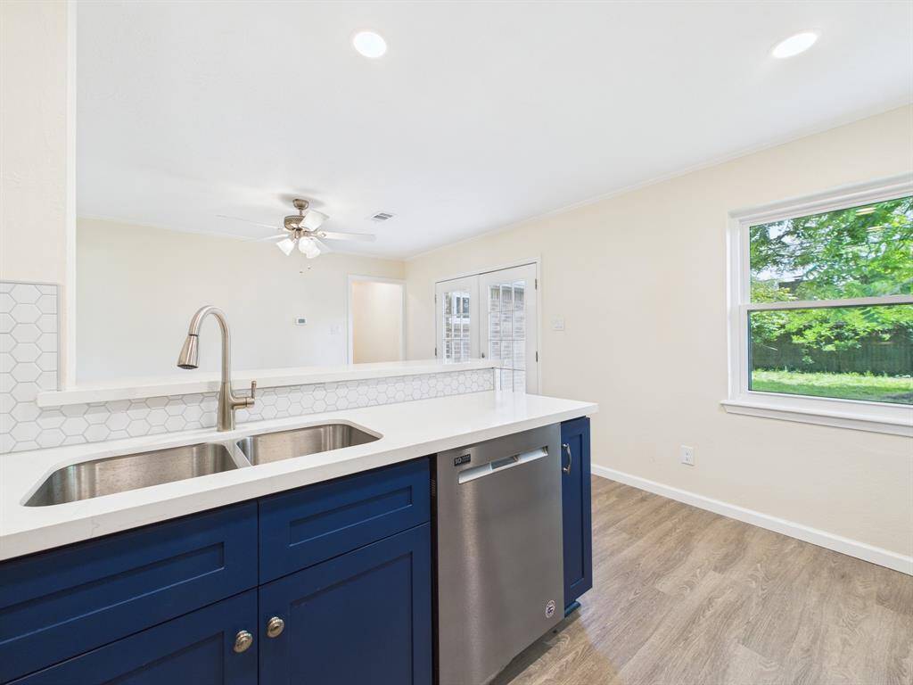 Bright kitchen with a modern design features a deep blue cabinet, stainless steel sink and dishwasher, and a large window offering a view of the lush backyard.