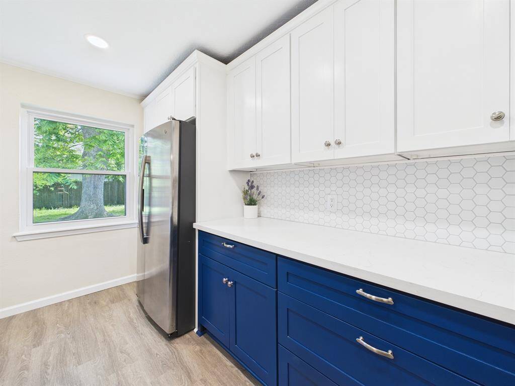 Modern kitchen featuring blue lower cabinets and white upper cabinets, with a sleek stainless steel fridge. The white hexagonal tile backsplash adds a stylish touch, and a large window provides plenty of natural light.