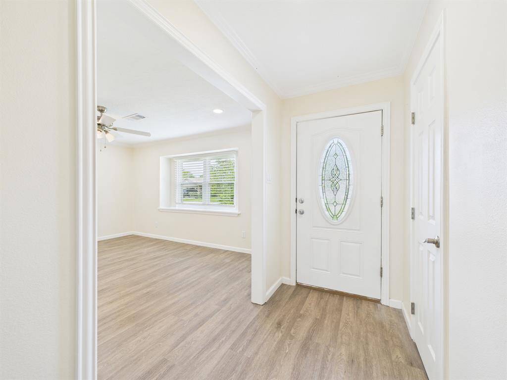 Bright entryway with a decorative glass front door and adjoining room view. Features light wood flooring, neutral walls, and abundant natural light from a large window.