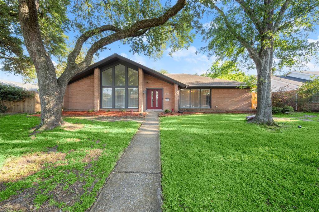 Charming brick home with a modern design, featuring large front windows and a welcoming red door. It's surrounded by a well-maintained lawn and mature trees, offering a serene and inviting atmosphere.