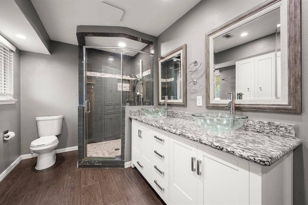 This modern bathroom features a sleek glass-enclosed shower, dual vessel sinks with granite countertops, and stylish framed mirrors. The space is enhanced by elegant wood-look flooring and a neutral color palette.
