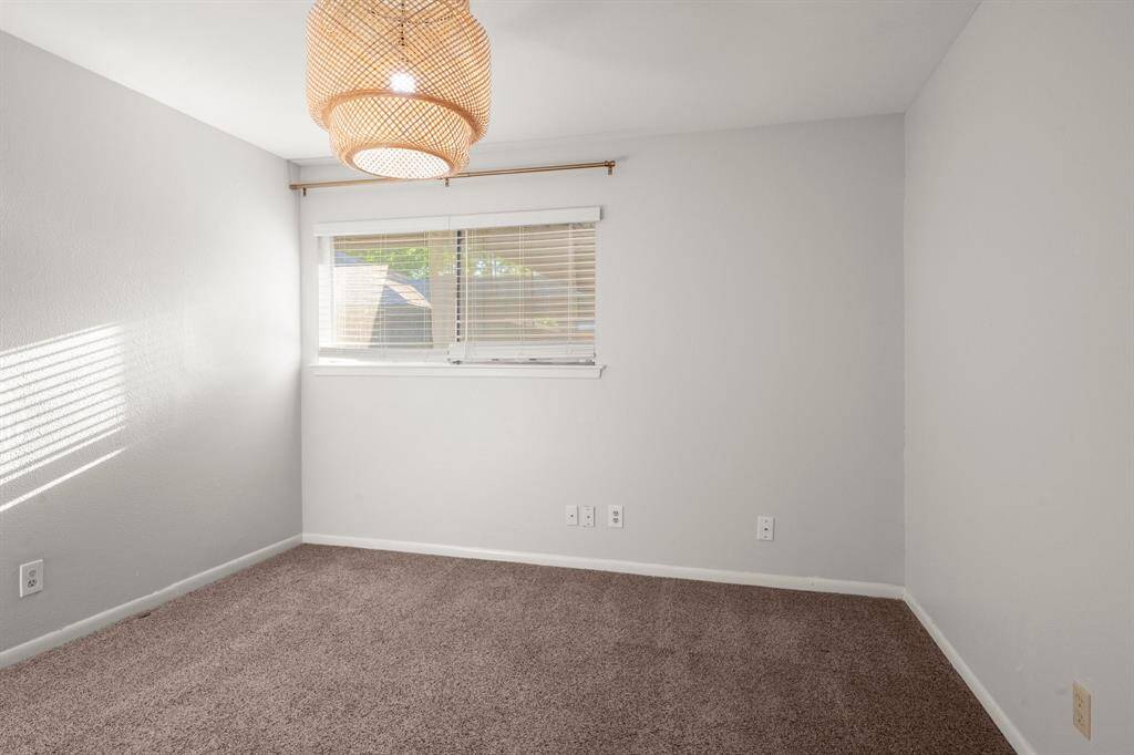 This room is cozy with neutral walls and carpet, featuring a large window with blinds that lets in natural light. A stylish woven pendant light adds a touch of elegance.