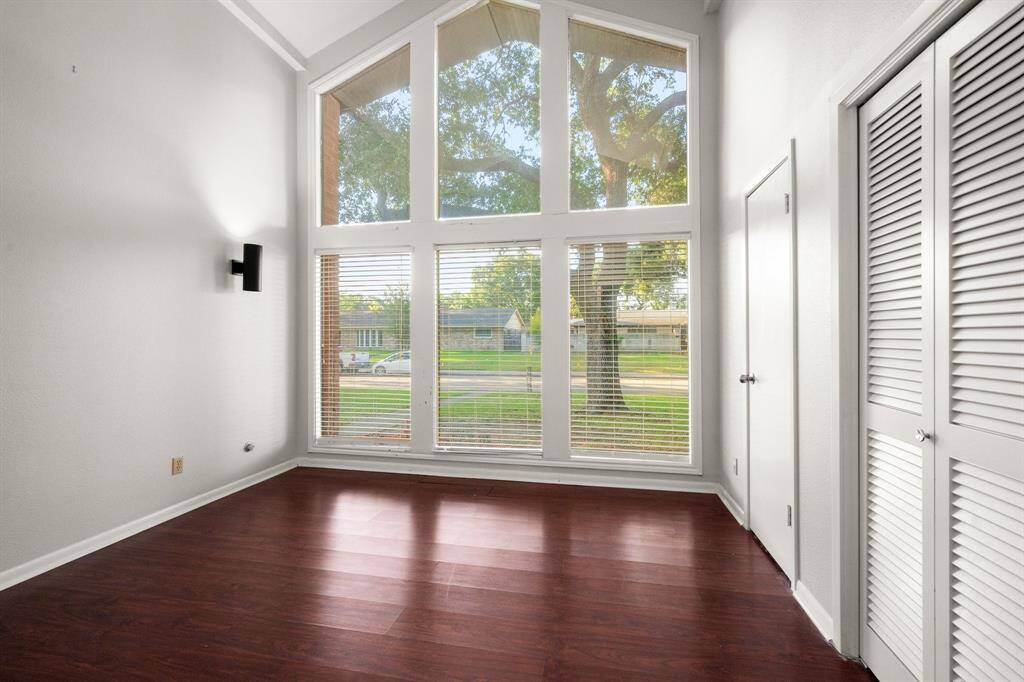 This room features high ceilings and large windows that provide ample natural light and a view of the greenery outside. It has hardwood floors and a modern wall sconce, with white walls and a closet with louvered doors.