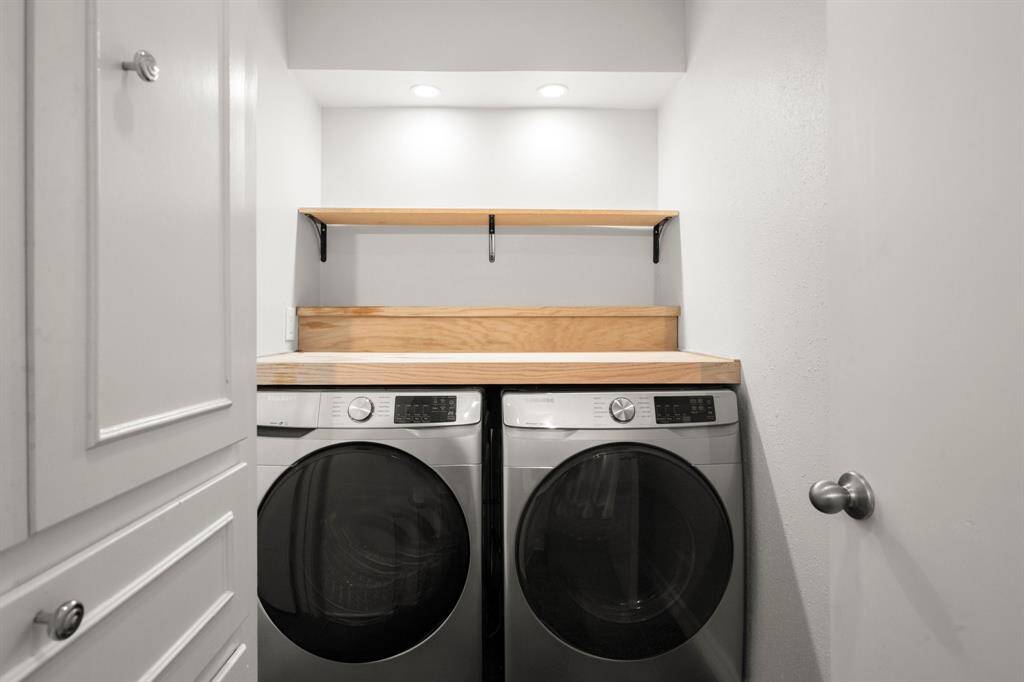 This photo shows a compact laundry area featuring modern front-loading washer and dryer units. It includes a wooden countertop and shelf for storage, with recessed lighting enhancing the clean, minimalistic design.