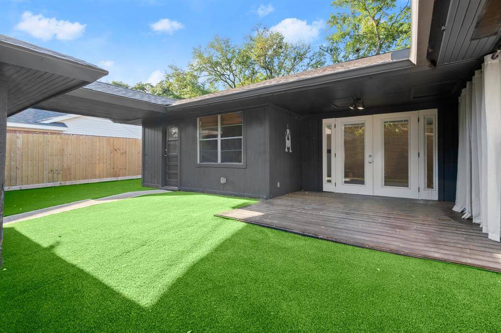 The photo showcases a modern, low-maintenance backyard featuring artificial grass and a wooden deck. The space is partially covered, with French doors leading indoors, offering a seamless transition for outdoor living. A wooden fence provides privacy, and the overall design emphasizes easy upkeep and functionality.