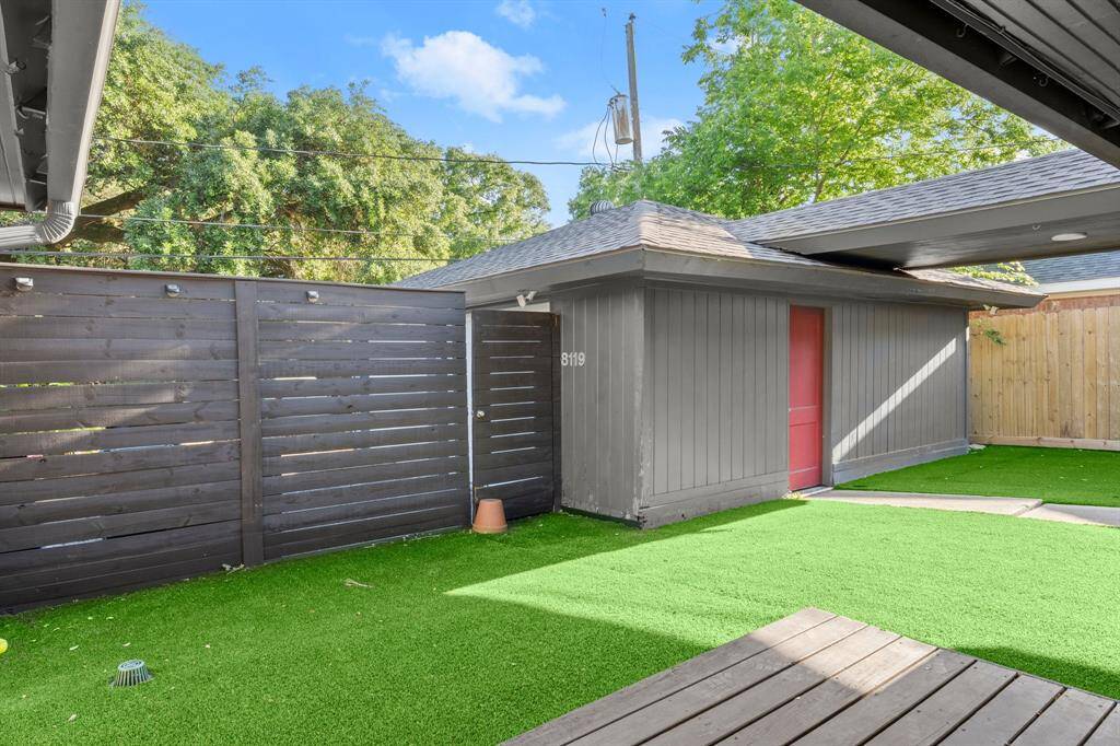 This photo shows a modern backyard with artificial grass and a wooden deck. It features a sleek, dark wood fence and a storage shed with a red door, creating a clean, low-maintenance outdoor space.