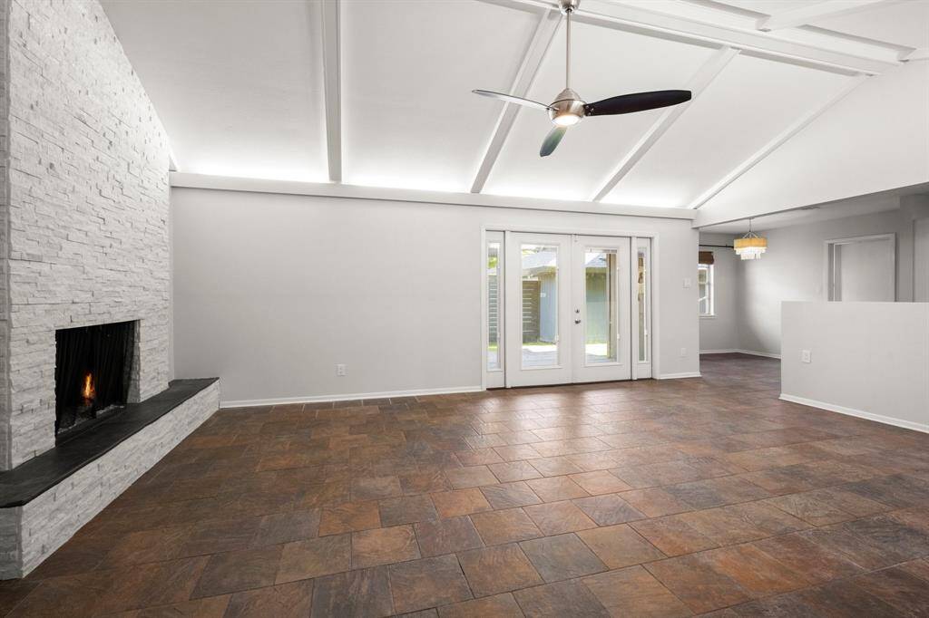 This spacious living area features a vaulted ceiling, a modern ceiling fan, and a stone fireplace. Natural light pours through the French doors, leading to the outdoor space. The room has neutral walls and tile flooring, creating a warm and inviting atmosphere.