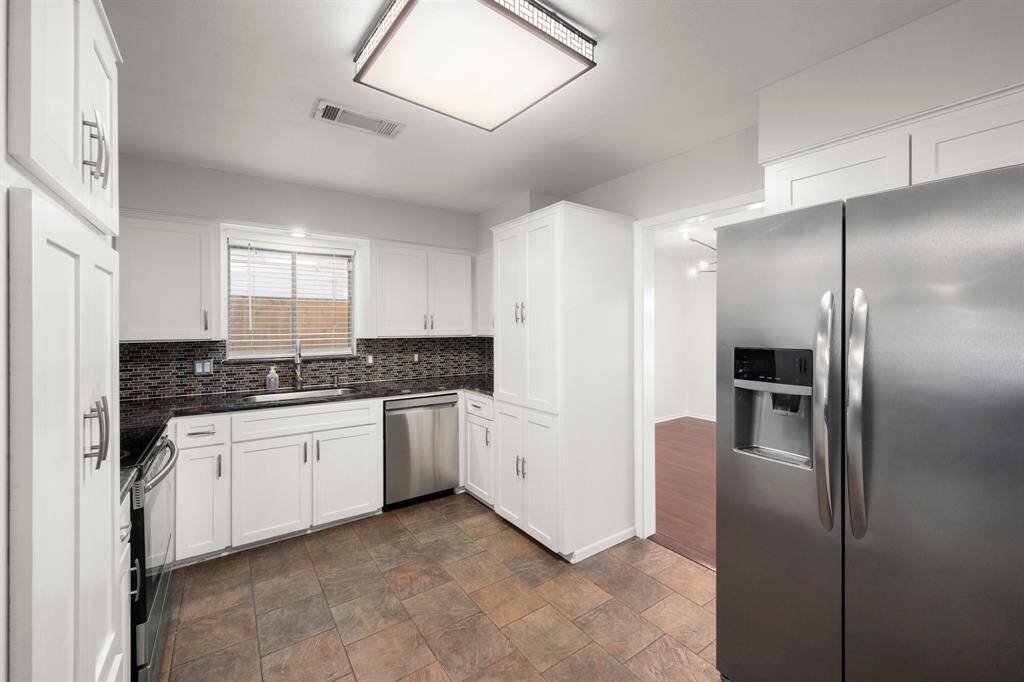 another view of the kitchen features modern stainless steel appliances, white cabinetry, and a sleek tile backsplash. It has a spacious layout with ample storage and a large window for natural light.