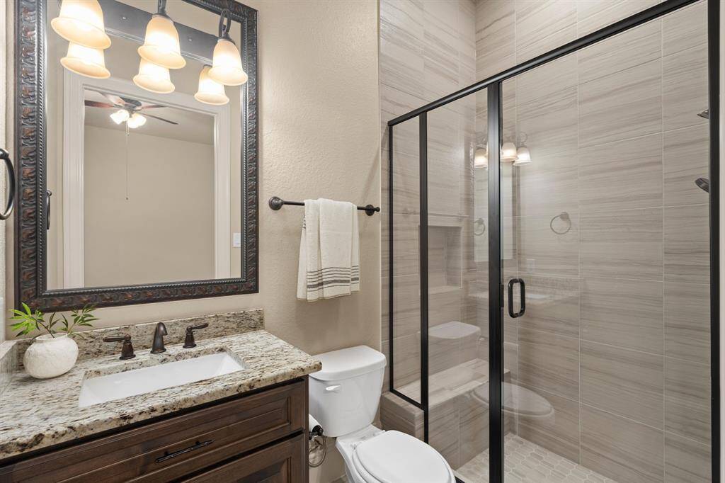 This spotless ensuite bathroom features a single sink granite vanity with a framed mirror and stylish vanity lights above. The convenient walk-in shower showcases beautiful tile work, a built-in bench, and a shampoo ledge, combining both elegance and functionality.