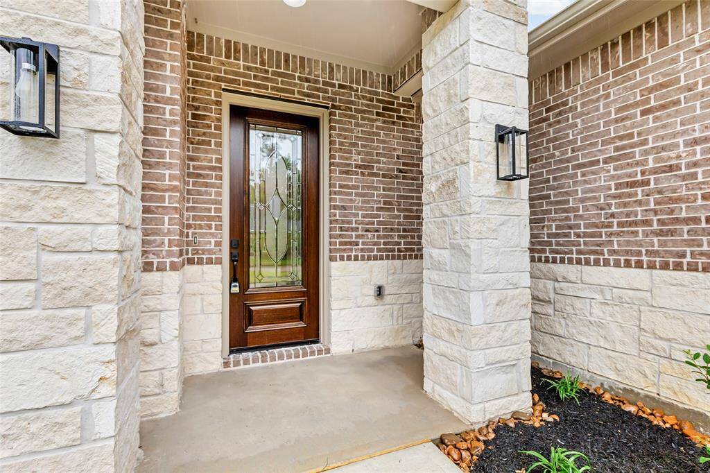 This welcoming entryway features a stylish wooden door with decorative glass, framed by a mix of brick and stone masonry. The porch is enhanced with modern wall-mounted lights and simple landscaping, creating a warm ambiance.