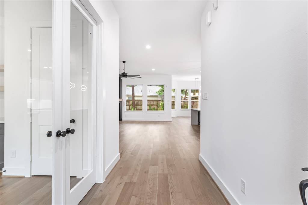 Bright, open-concept hallway with hardwood floors and white walls. There's a glass door on the left leading to the office/4th bedroom, and large windows at the end provide natural light and a view of the fenced backyard.