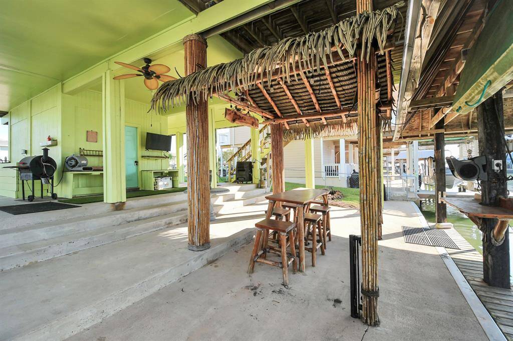 A tropical-themed patio area with a thatched-roof overhang, a wooden bar table, and stools. It features a vibrant color scheme, ceiling fan, grill, situated next to a boat dock, ideal for outdoor entertaining.