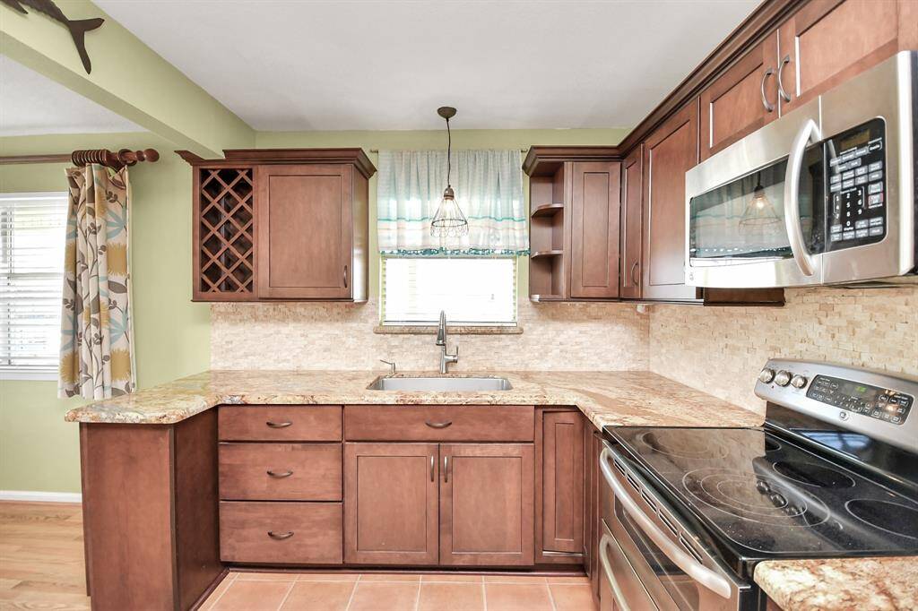 This kitchen features rich wooden cabinetry with a built-in wine rack, granite countertops, and a stone tile backsplash. It's equipped with stainless steel appliances, including a microwave and stove. A window above the sink provides natural light, and the space is accented by light green walls.