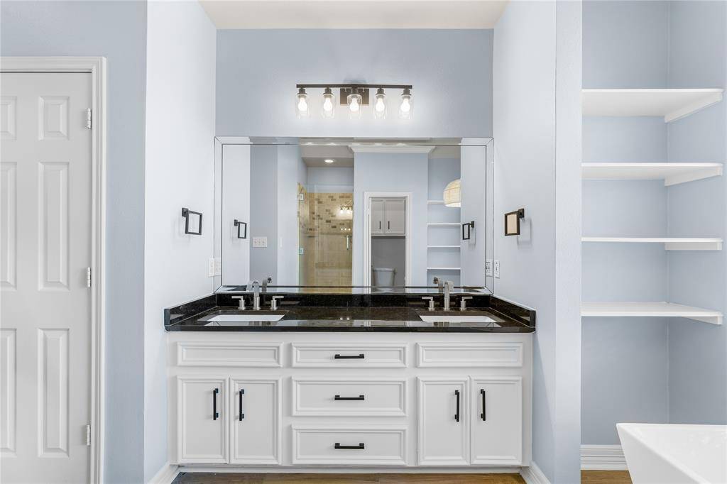 Primary bath with granite countertops, updated lighting and hardware, plus ample cabinet space. It features built-in shelves for storage and a large mirror with overhead lighting & wood floors.