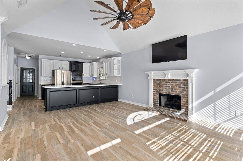 The den offers a bright open-plan living area with a modern kitchen featuring stainless steel appliances, white cabinetry, and a contrasting island. The room includes a cozy fireplace and is well-lit with natural light.