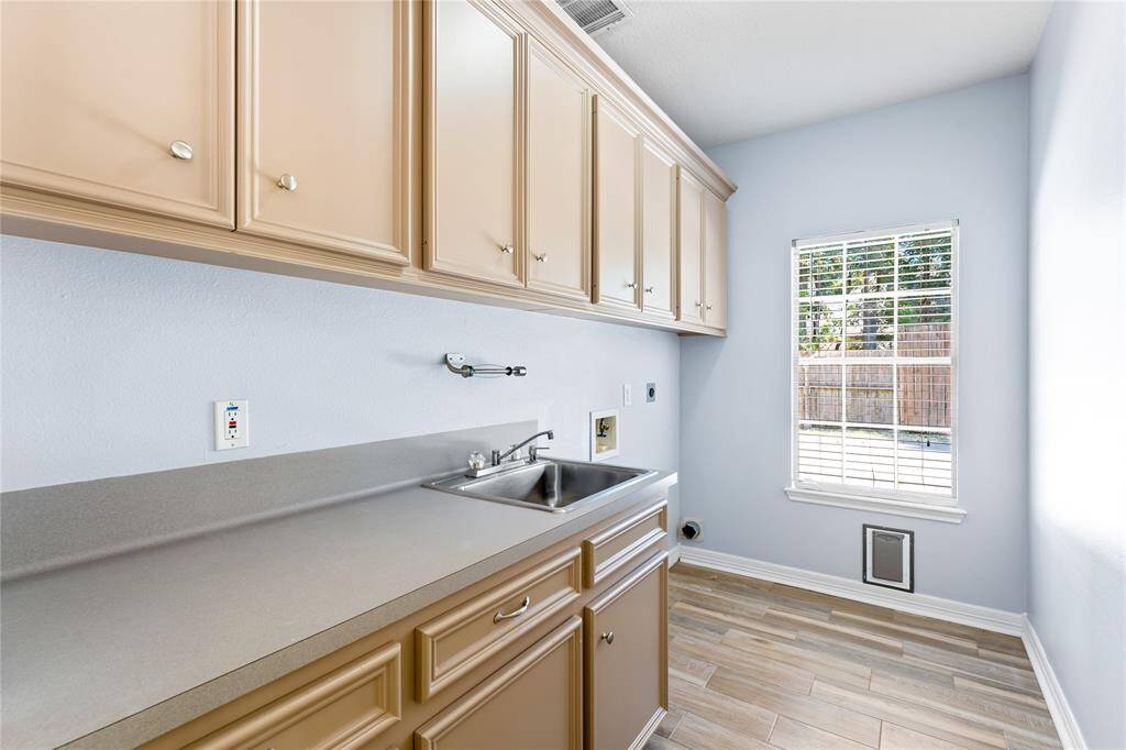 Oversized laundry room featuring room for a fridge, upper & lower cabinets, sink, neutral countertops, and light wood flooring. A window allows for natural light.