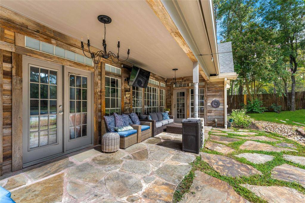 Another view of the covered back patio with rustic charm, featuring French doors, comfortable seating, and a natural stone floor, all overlooking a peaceful, tree-lined backyard with pool and spa.