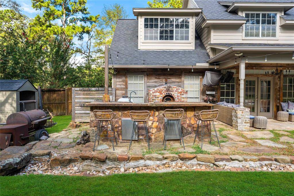 This is an inviting backyard with a covered patio, brick pizza oven, outdoor bar seating, and a barbecue setup, ideal for outdoor entertaining within a fenced, private space surrounded by greenery.
